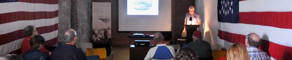 A man stands behind a podium while giving a presentation on a screen while a small audience looks on.