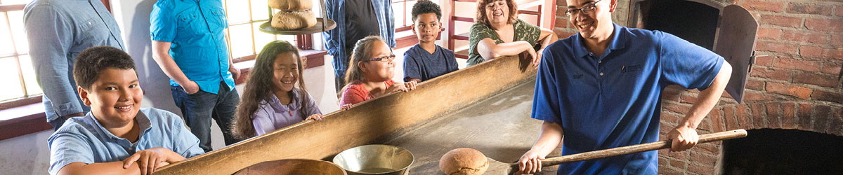 A Fort Snelling staff person baking bread in a historic oven as visitors watch.