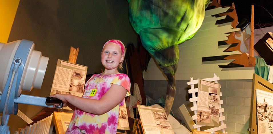A girl holding a directional fan standing in front of a cloth tornado and damaged house.