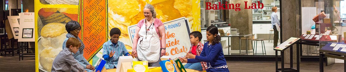 A group of children interact with a museum exhibit while a volunteer looks on.