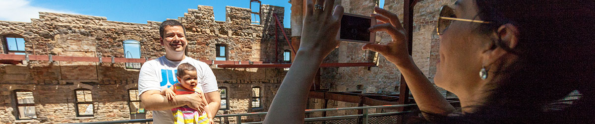 A woman stands back to take a photo of a man with a baby in front of the ruin courtyard.
