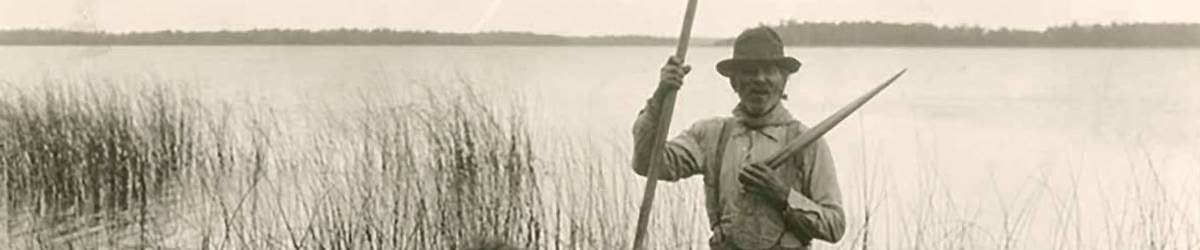 A man in a hat holding a boat paddle standing near a lake with wild rice growing.