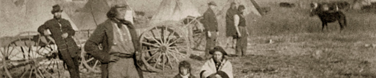 Two people kneeling while several people standing with many tipi in the background.