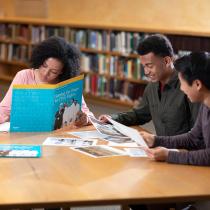 Three teenagers sit at a table looking at papers and writing notes.